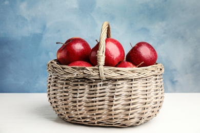 Photo of Wicker basket with ripe juicy red apples on white wooden table against blue background