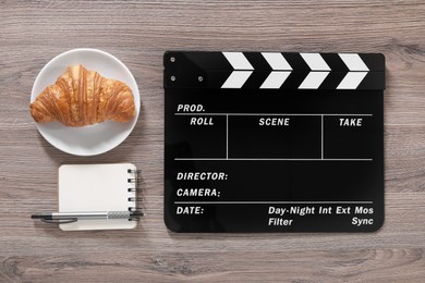 Photo of Movie clapper, croissant, notebook and pen on wooden table, flat lay