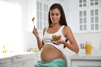Photo of Young pregnant woman with bowl of vegetable salad at table in kitchen. Taking care of baby health