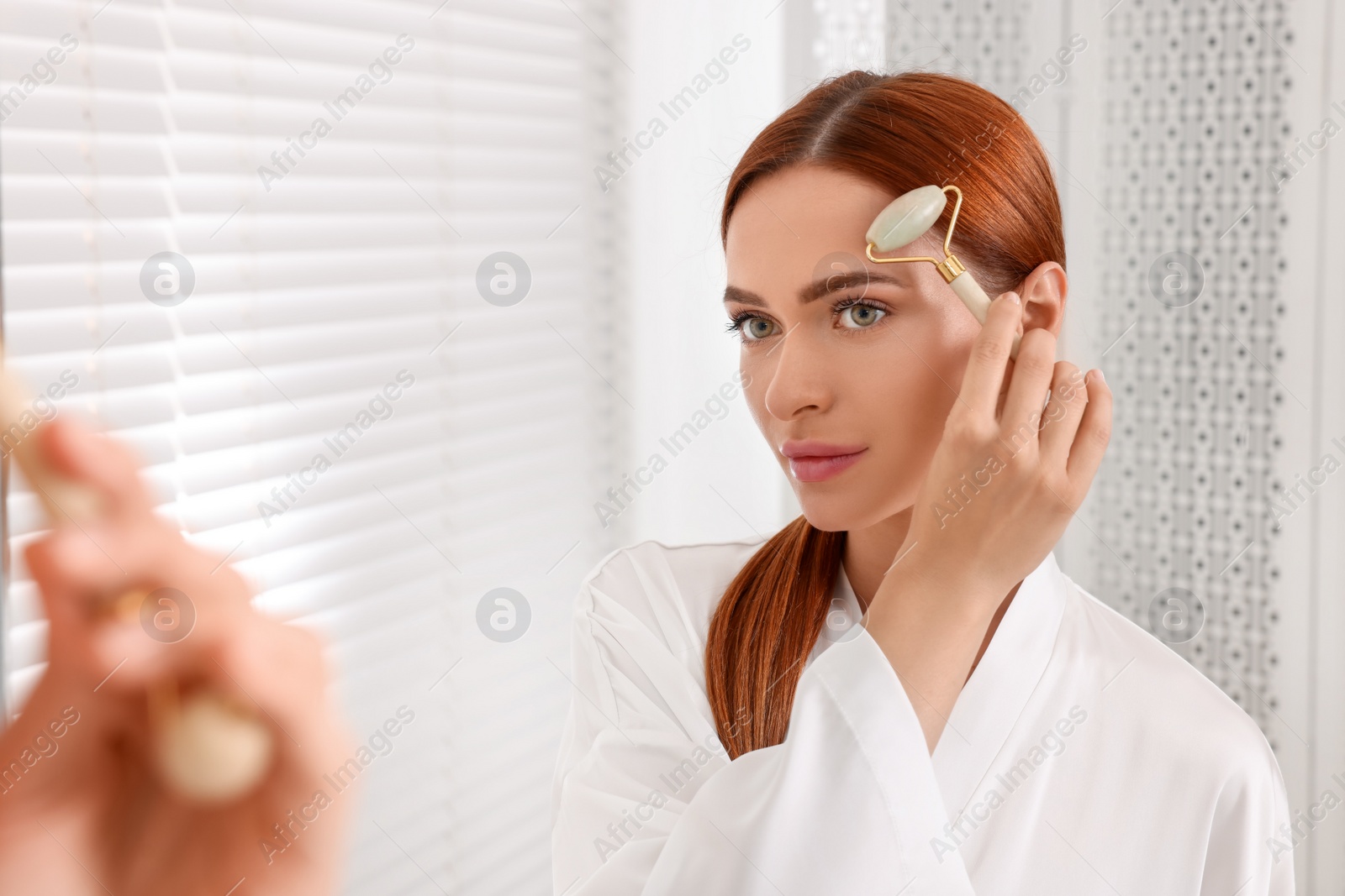 Photo of Young woman massaging her face with jade roller near mirror in bathroom