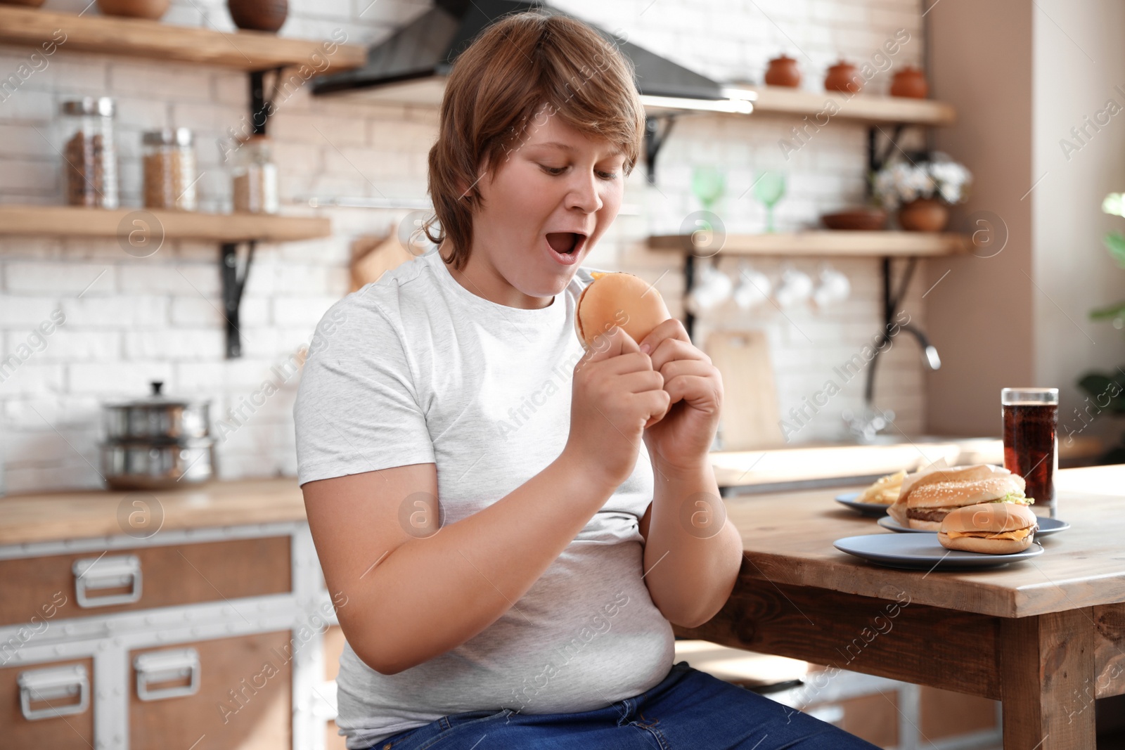 Photo of Emotional overweight boy at table with fast food in kitchen