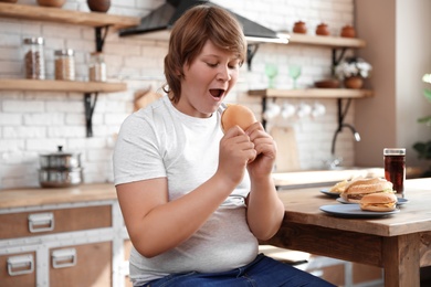 Photo of Emotional overweight boy at table with fast food in kitchen