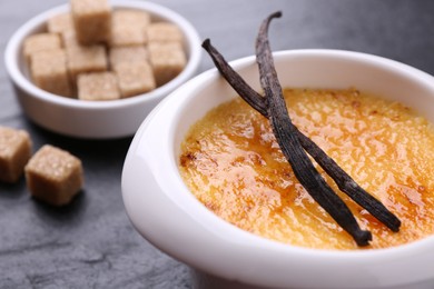 Delicious creme brulee in bowl, vanilla pods and sugar cubes on dark gray table, closeup