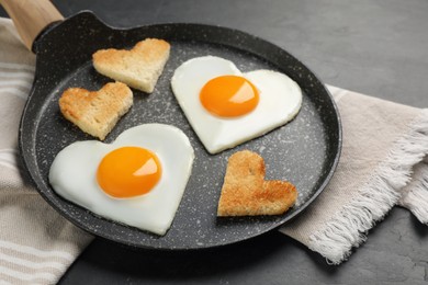Photo of Heart shaped fried eggs and toasts in frying pan on grey table, closeup