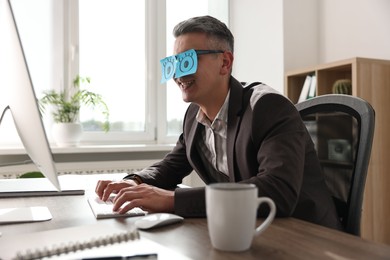 Photo of Man with fake eyes painted on sticky notes working on computer at table in office