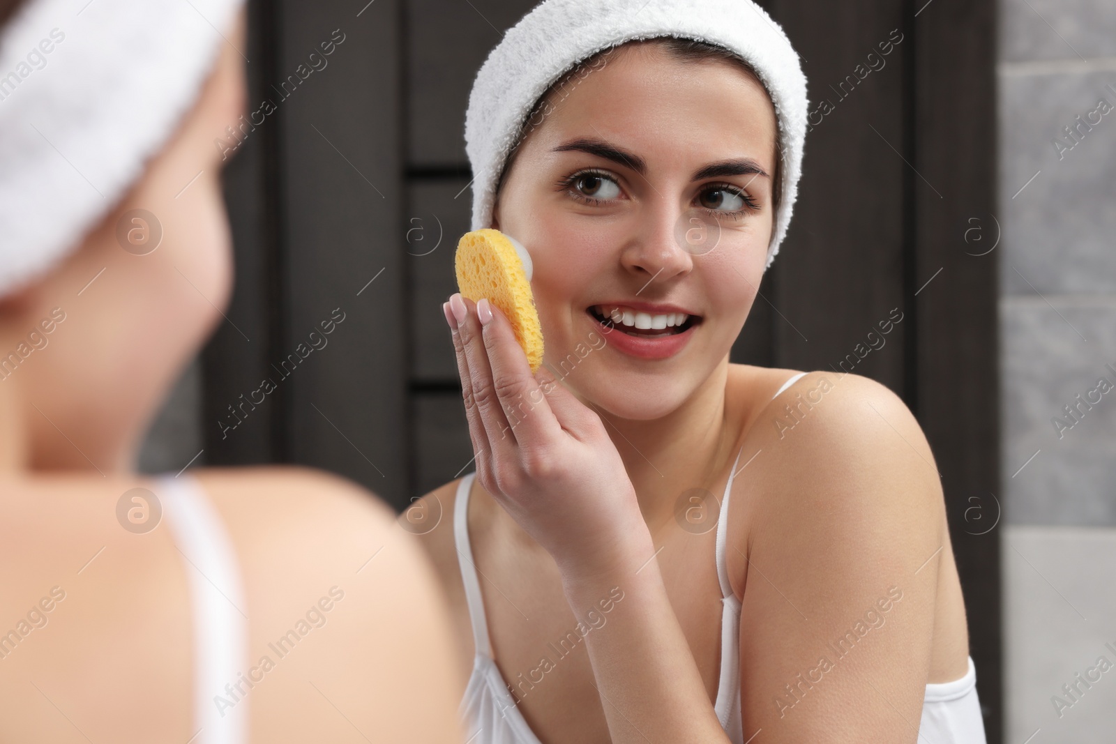 Photo of Young woman with headband washing her face using sponge near mirror in bathroom