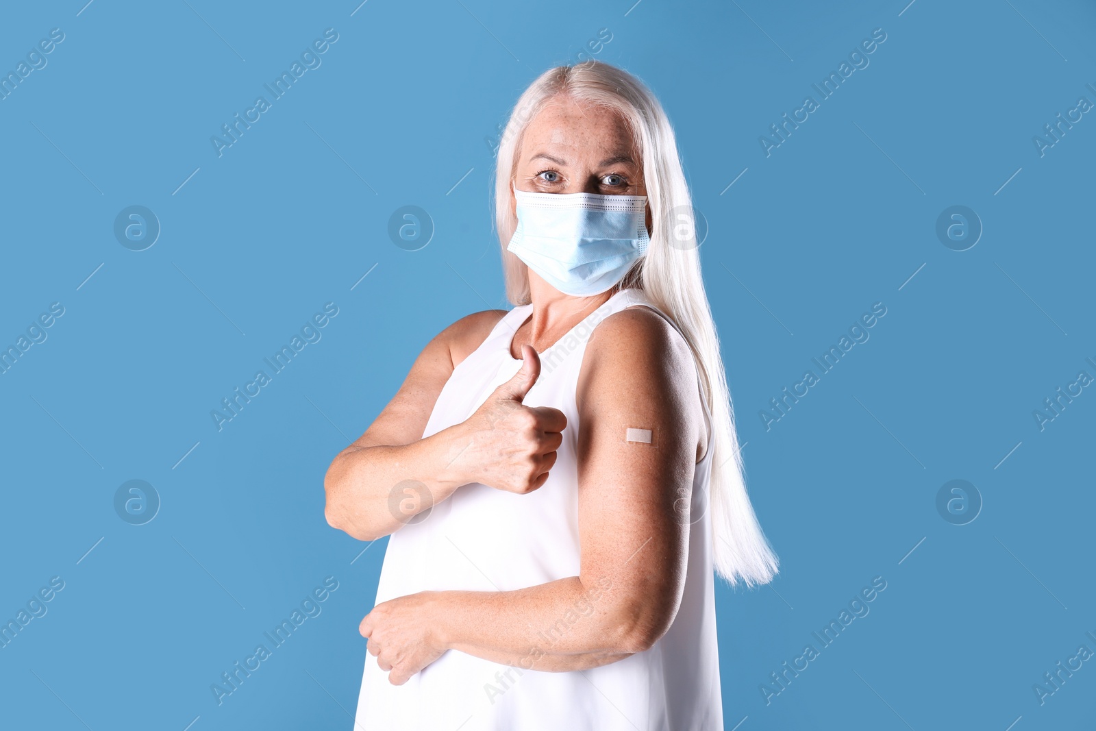 Photo of Mature woman in protective mask showing arm with bandage after vaccination and thumb up on light blue background