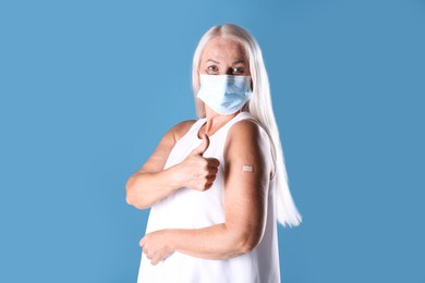 Mature woman in protective mask showing arm with bandage after vaccination and thumb up on light blue background