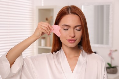 Photo of Young woman massaging her face with rose quartz gua sha tool in bathroom