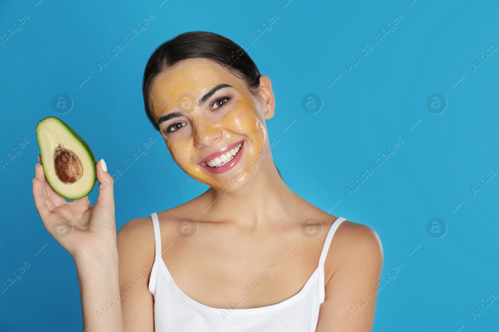 Photo of Young woman with cleansing mask on her face holding avocado against color background, space for text. Skin care