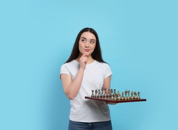Thoughtful woman holding chessboard with game pieces on light blue background, space for text