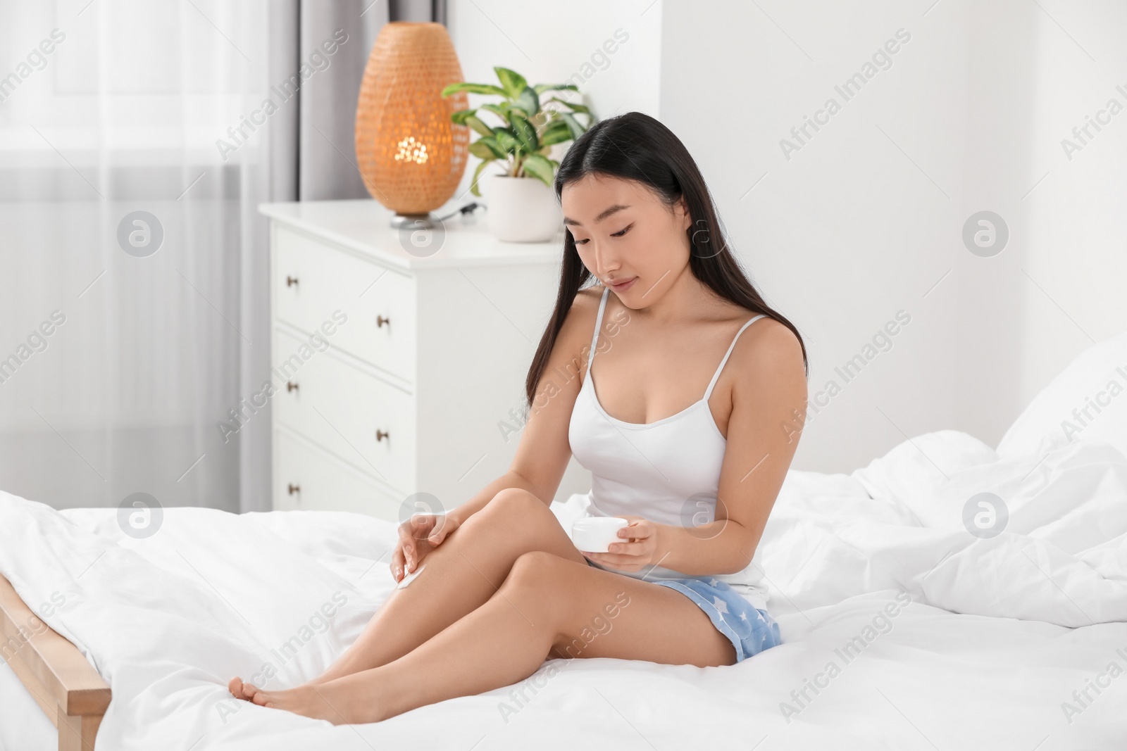 Photo of Beautiful young Asian woman applying body cream on leg in bedroom