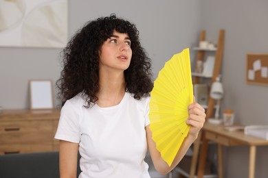 Photo of Young woman waving yellow hand fan to cool herself at home