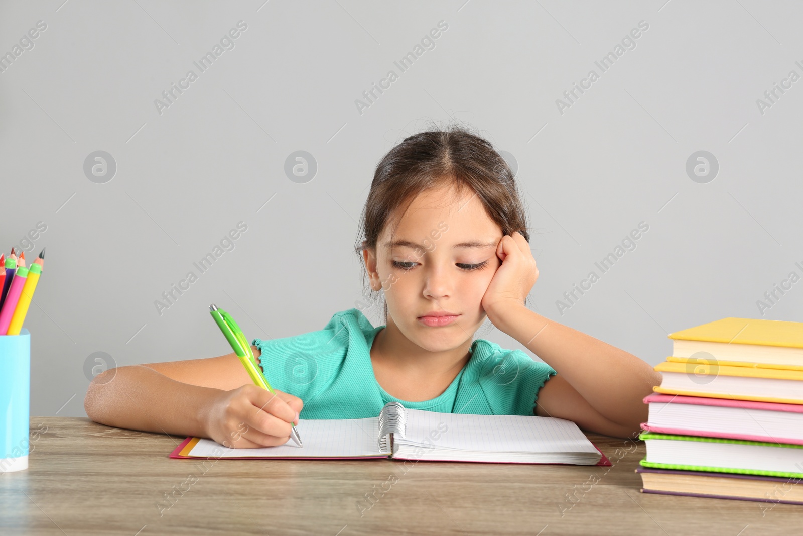 Photo of Bored little girl doing homework at table on grey background