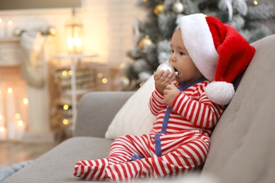 Image of Cute baby in Santa hat sitting on sofa at home. Christmas celebration