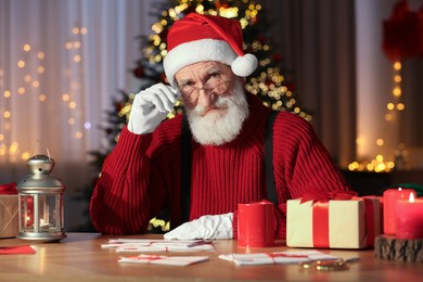 Photo of Santa Claus at his workplace. Letters, cup of beverage and gift boxes on table in room with Christmas tree