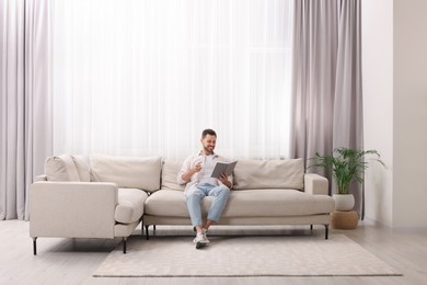 Photo of Happy man reading book on sofa near window with beautiful curtains in living room