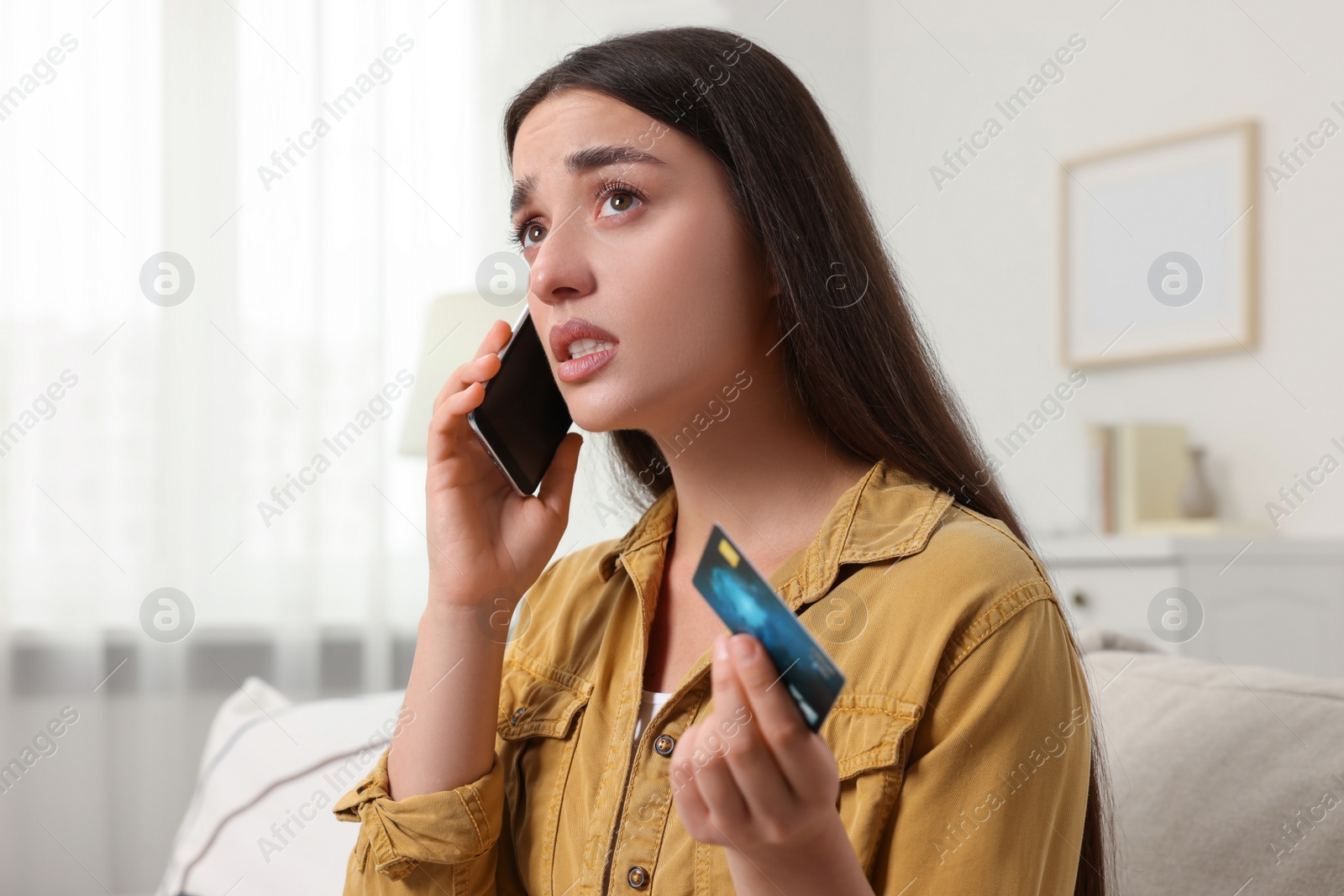 Photo of Confused woman with credit card talking on phone at home. Be careful - fraud