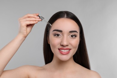 Photo of Happy young woman applying essential oil onto face on light grey background