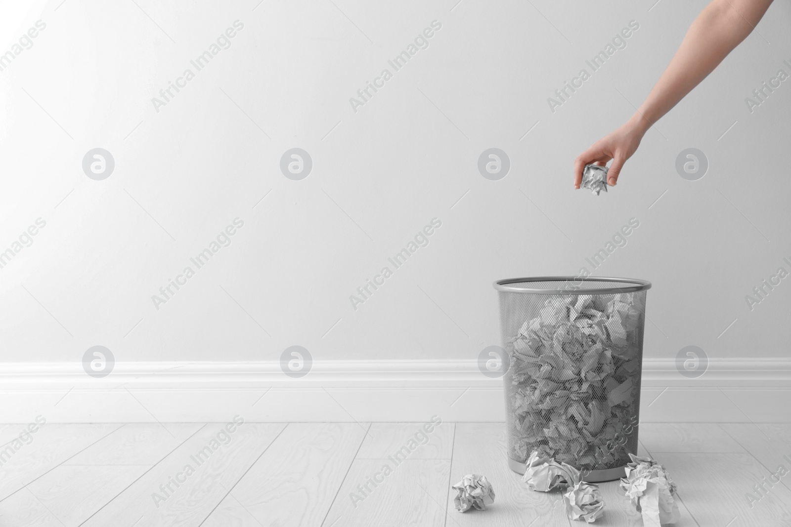 Photo of Woman throwing crumpled paper into metal bin against light wall. Space for text