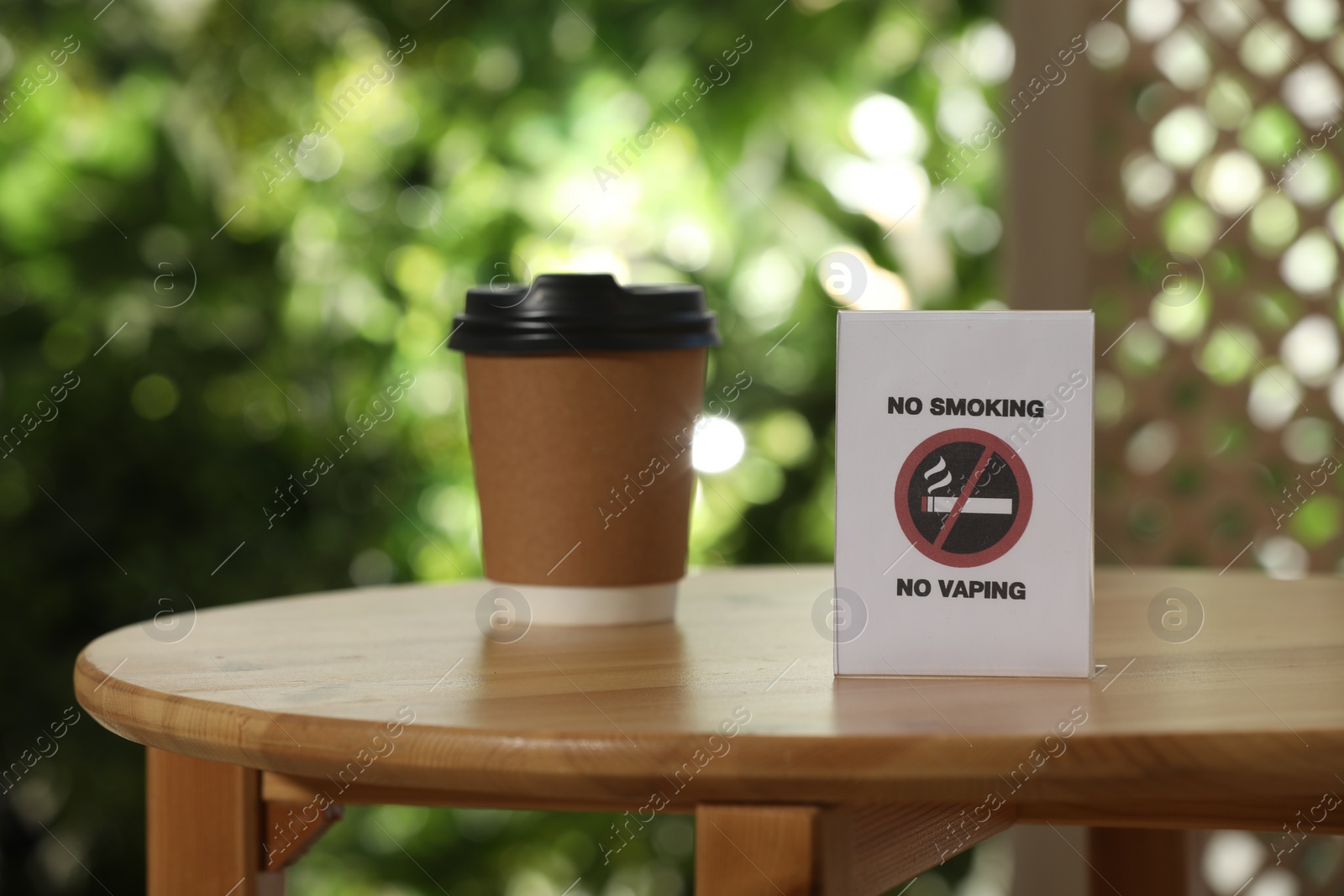 Photo of No Smoking No Vaping sign and cup of drink on wooden table outdoors, selective focus