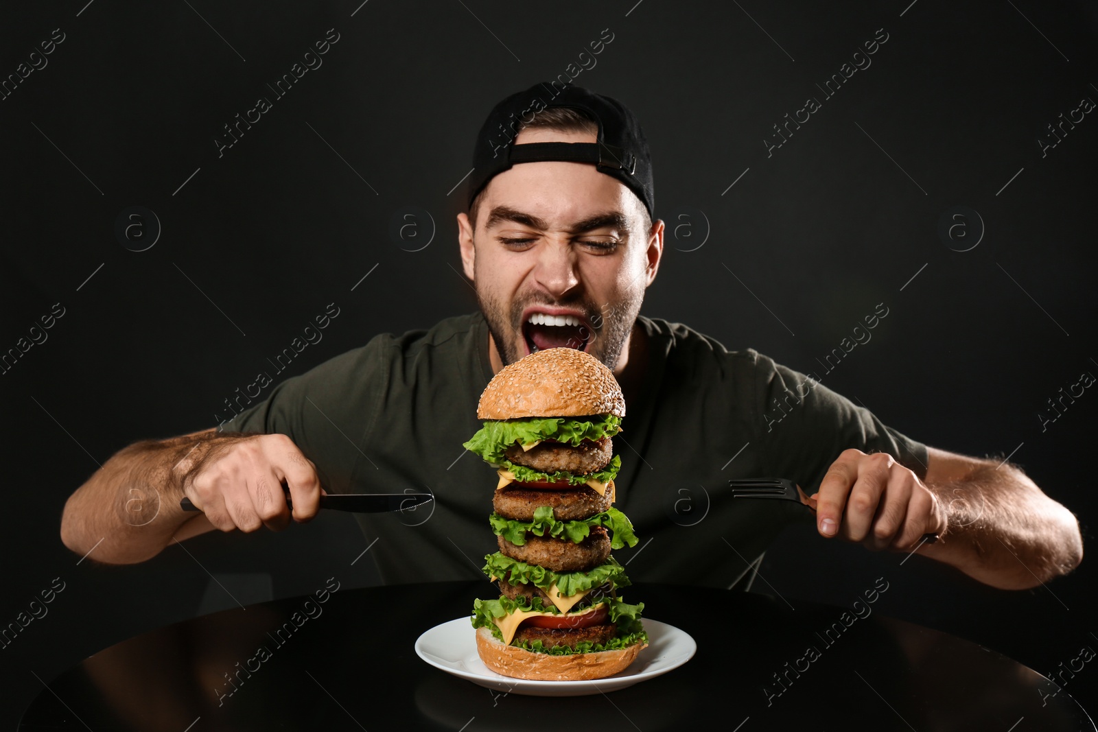 Photo of Young hungry man with cutlery eating huge burger on black background