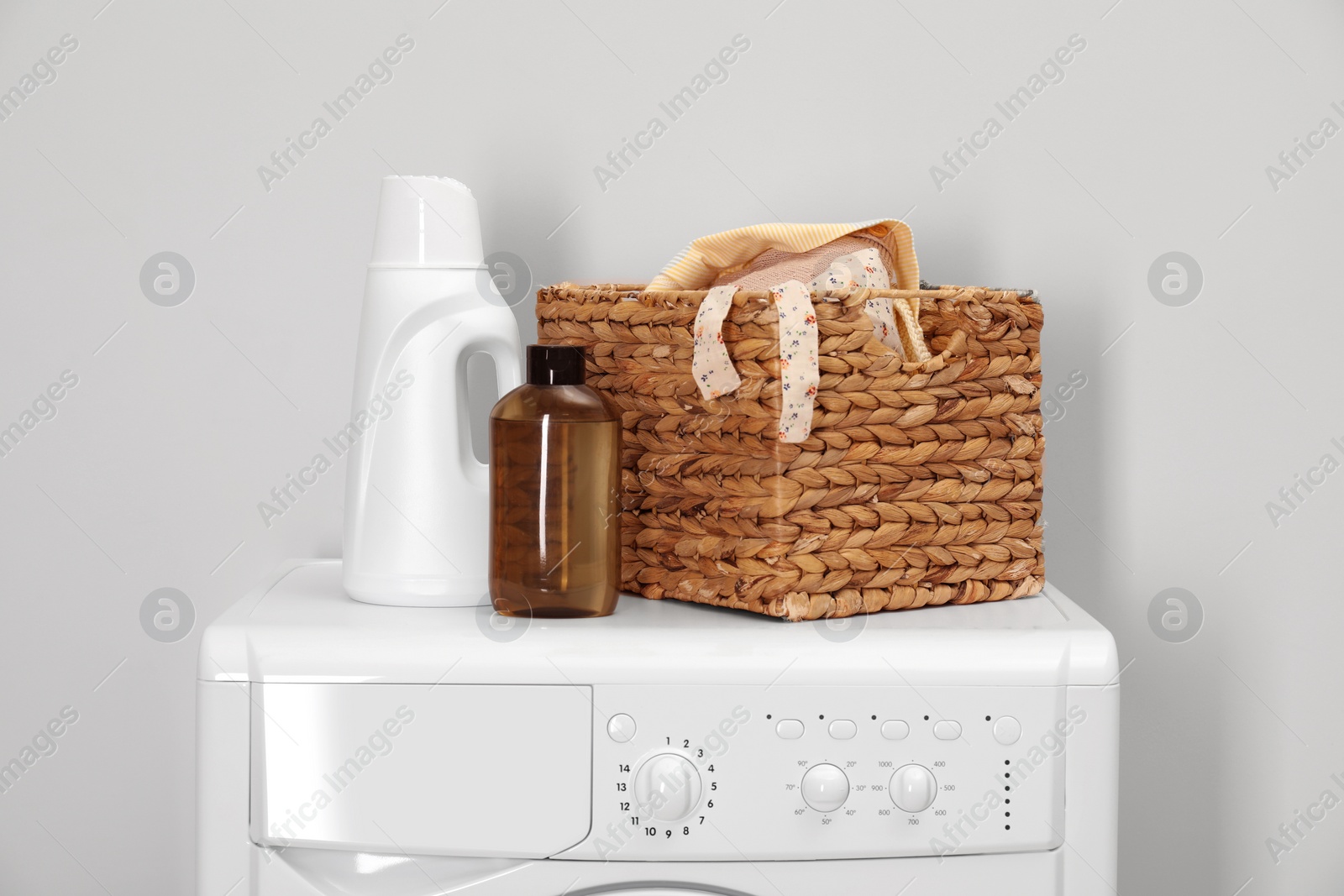 Photo of Baby clothes in wicker basket and laundry detergents on washing machine near light wall