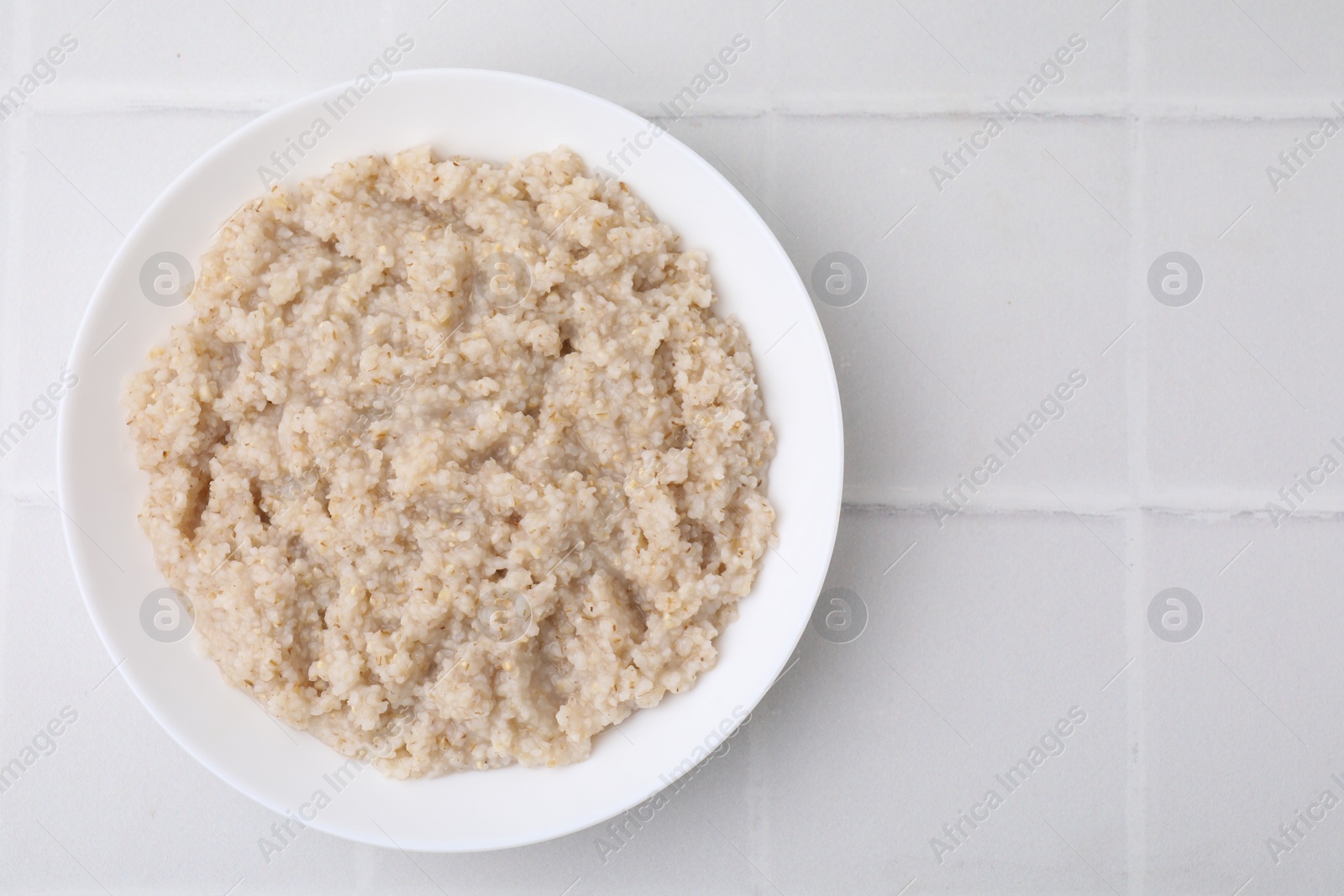 Photo of Delicious barley porridge in bowl on white tiled table, top view. Space for text