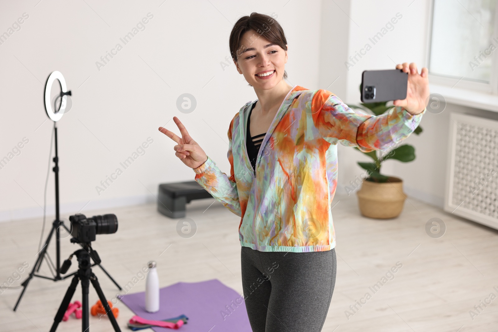 Photo of Happy sports blogger showing peace sign while streaming online fitness lesson with smartphone at home