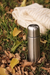 Photo of Metallic thermos and knitted scarf on green grass covered with fallen leaves outdoors