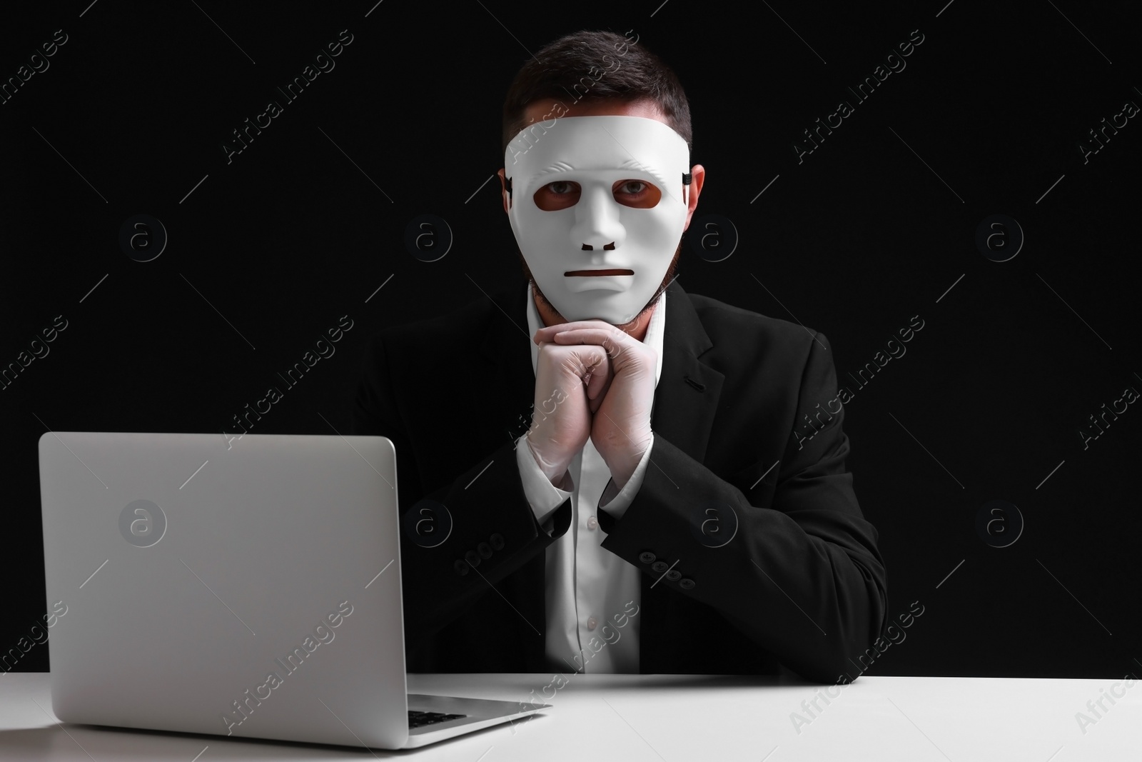 Photo of Man in mask and gloves sitting with laptop at white table against black background