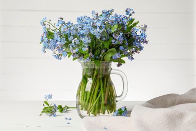 Photo of Bouquet of beautiful forget-me-not flowers in glass jug and blue cloth on white table