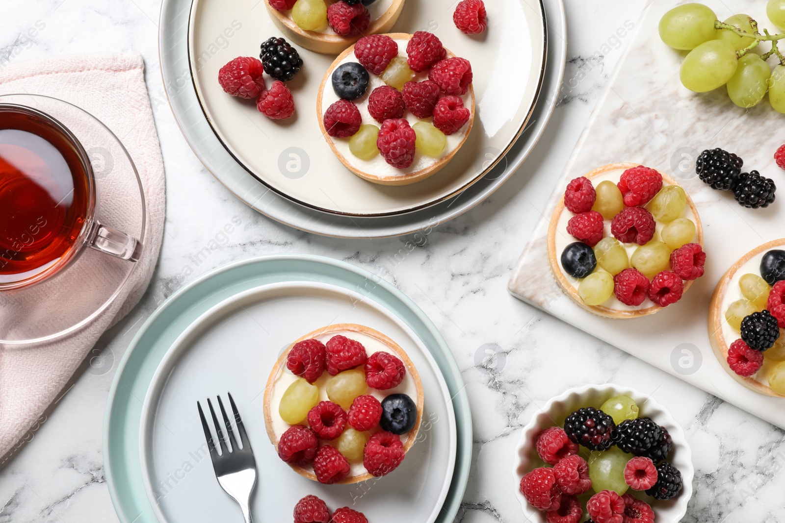 Photo of Delicious tartlets with berries and tea on white marble table, flat lay