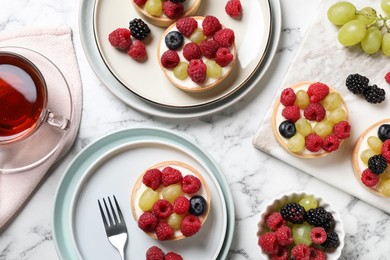 Delicious tartlets with berries and tea on white marble table, flat lay