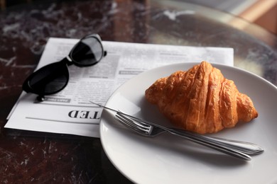 Tasty croissant, newspaper and sunglasses on black table