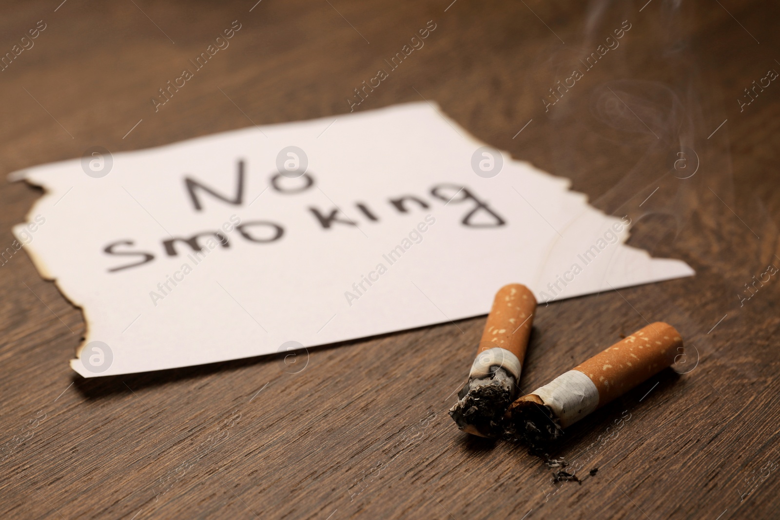 Photo of Burnt cigarettes and words No Smoking written on paper on wooden table, closeup