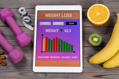 Photo of Tablet with weight loss calculator application, dumbbells and products on grey wooden table, above view