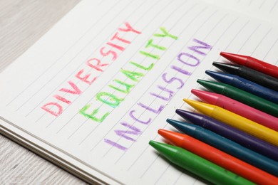 Photo of Notebook with words Diversity, Equality, Inclusion and wax pencils on wooden table, closeup