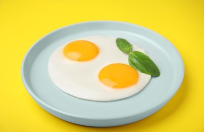 Tasty fried eggs with basil in plate on yellow background, closeup