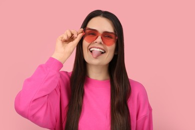 Photo of Happy young woman with heart shaped glasses showing her tongue on pink background
