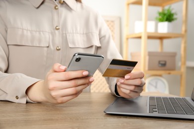 Online payment. Woman using smartphone and credit card near laptop at wooden table indoors, closeup