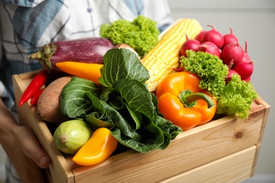 Woman holding wooden crate full of fresh vegetables on light background, closeup
