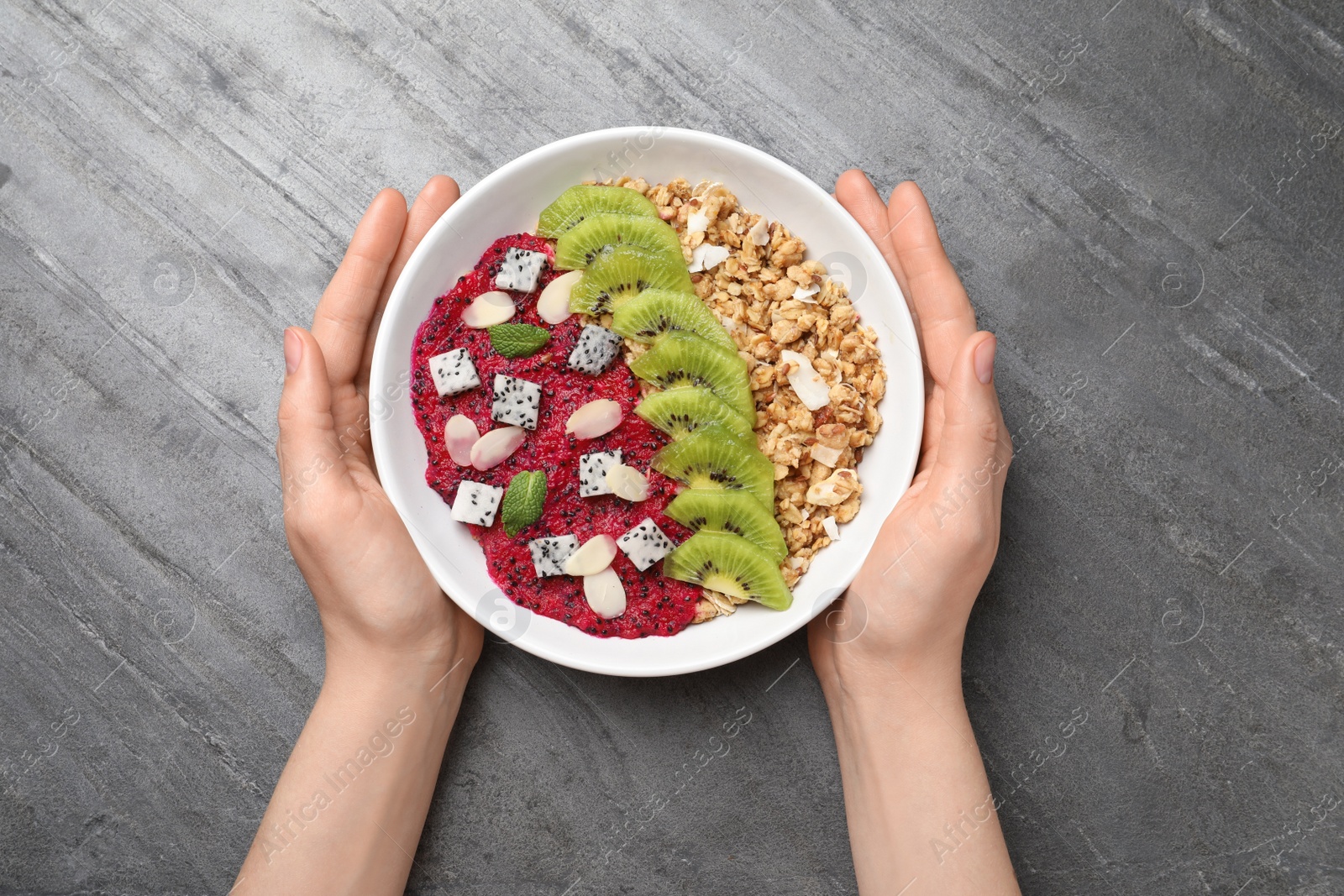 Photo of Woman holding bowl of granola with pitahaya, kiwi and almonds at grey table, top view