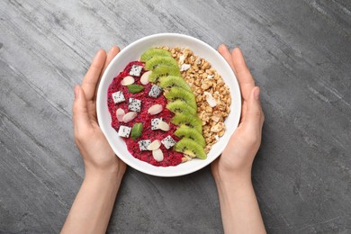 Photo of Woman holding bowl of granola with pitahaya, kiwi and almonds at grey table, top view