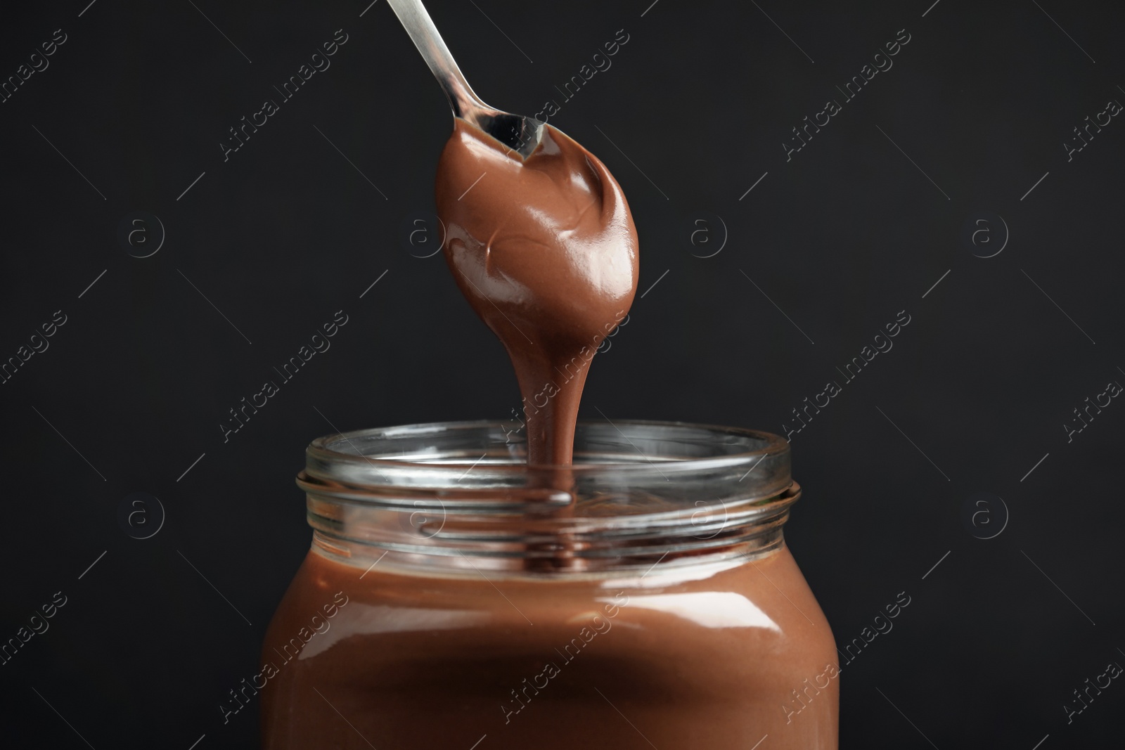 Photo of Pouring sweet chocolate cream into glass jar on dark background