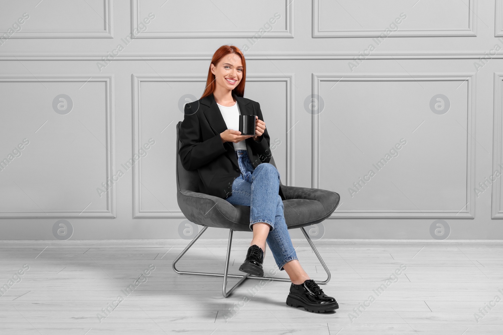 Photo of Happy young woman with cup of drink sitting in armchair near light grey wall indoors