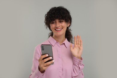 Happy young woman looking at smartphone and waving hello on light grey background