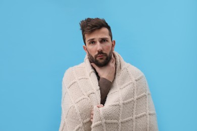 Photo of Man wrapped in blanket suffering from sore throat on light blue background. Cold symptoms