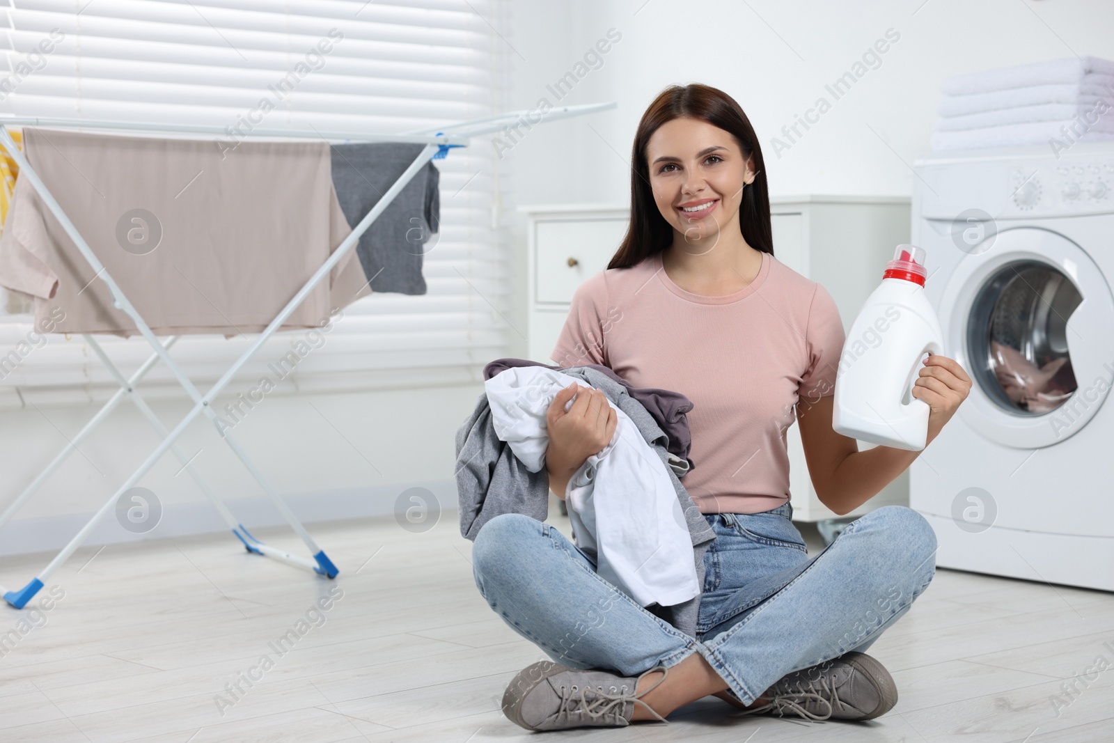 Photo of Woman sitting on floor near washing machine and holding fabric softener with dirty clothes in bathroom, space for text