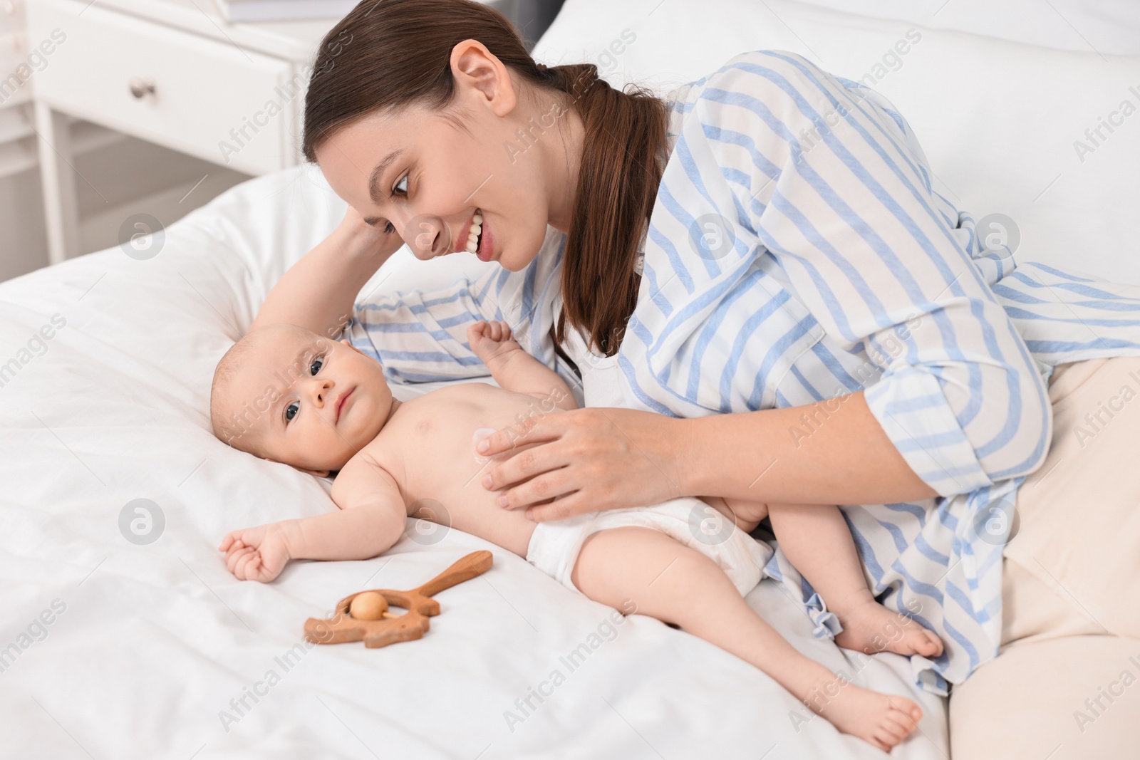 Photo of Happy young woman applying body cream onto baby`s skin on bed